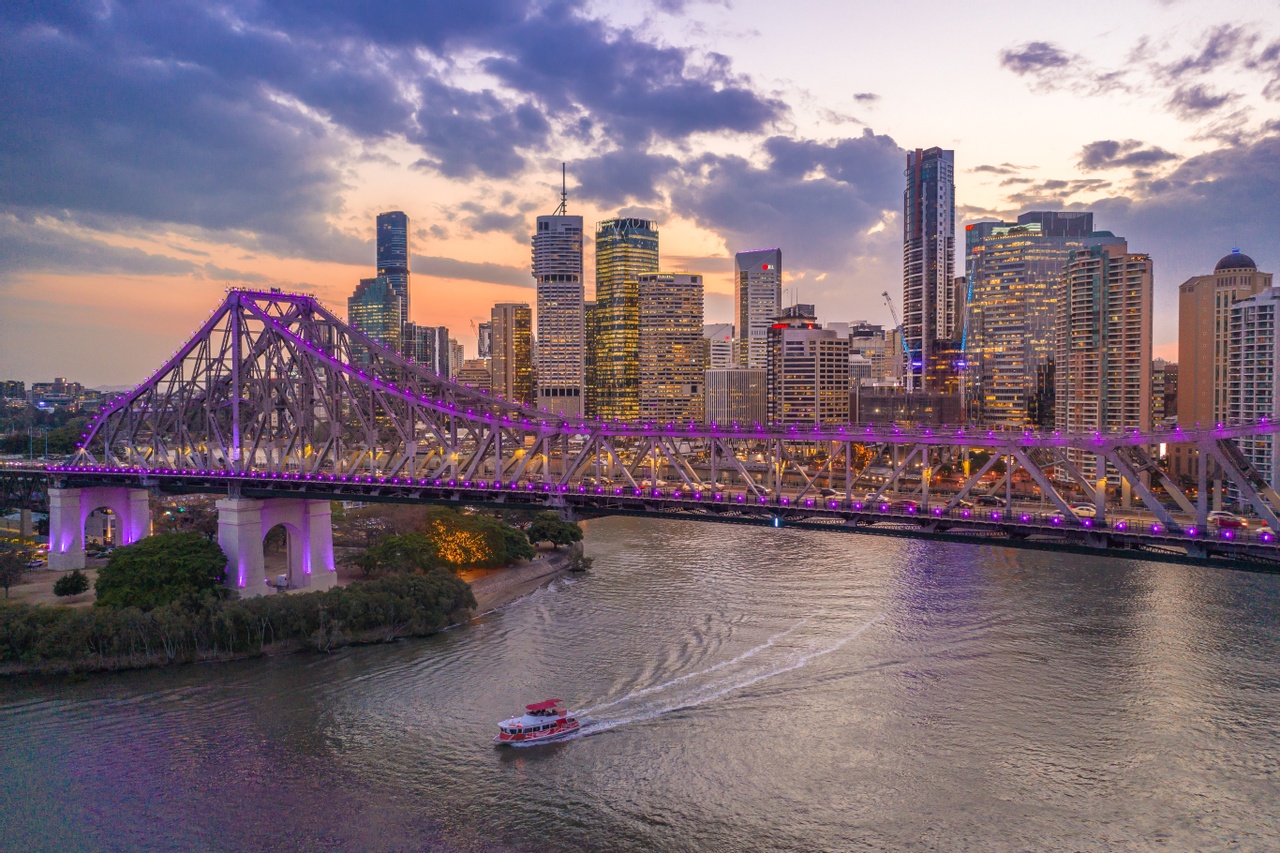 The Story Bridge lit up in Purple lights as the sun sets with city as background.