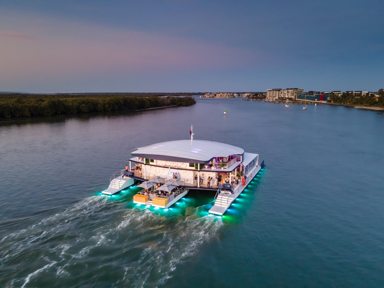 An image of a large YOT Club yacht with neon lights travelling down a river at dusk.