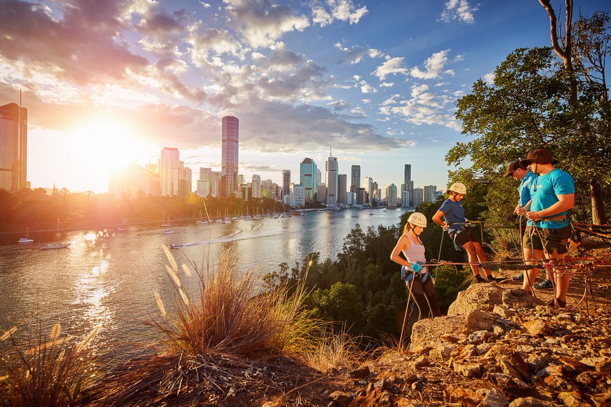 One female and one male climbers getting ready for cliff climbing game with two staff in blue shirts and Brisbane River sunset as background at Kangaroo Point Cliffs.