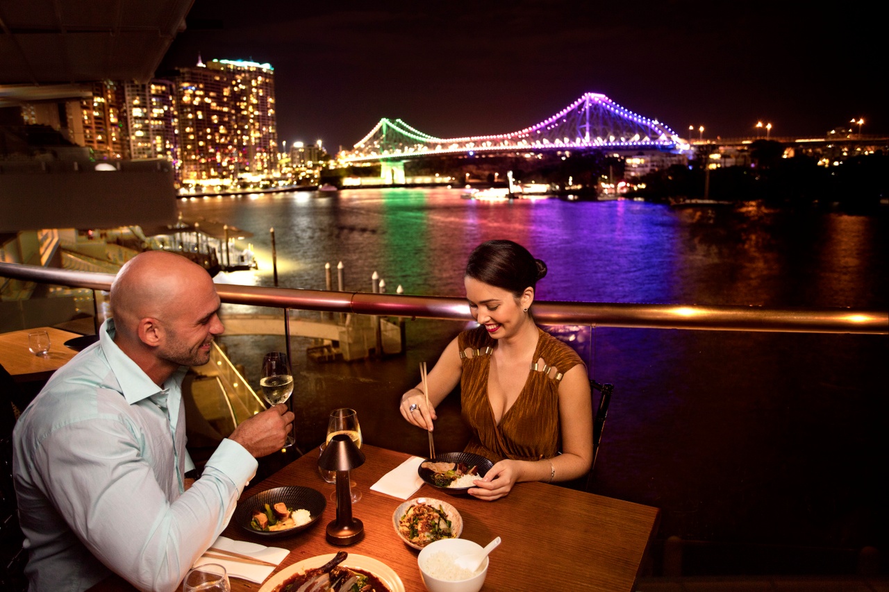 Two people dining by the riverside at night with colour lighting bridge as background.