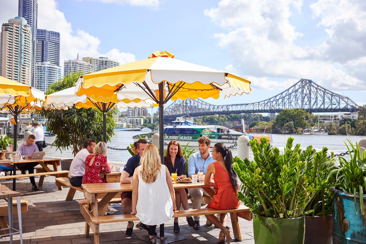 A group of people dining alfresco by the river under an umbrella at CBD in daytime.