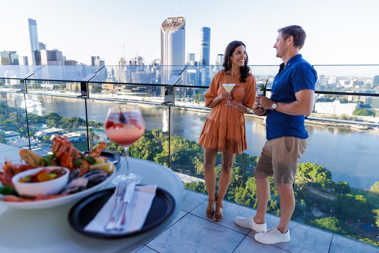 A couple talking on Emporium Hotel Terrace Rooftop Bar overlooking the river with seafood plater at the front.