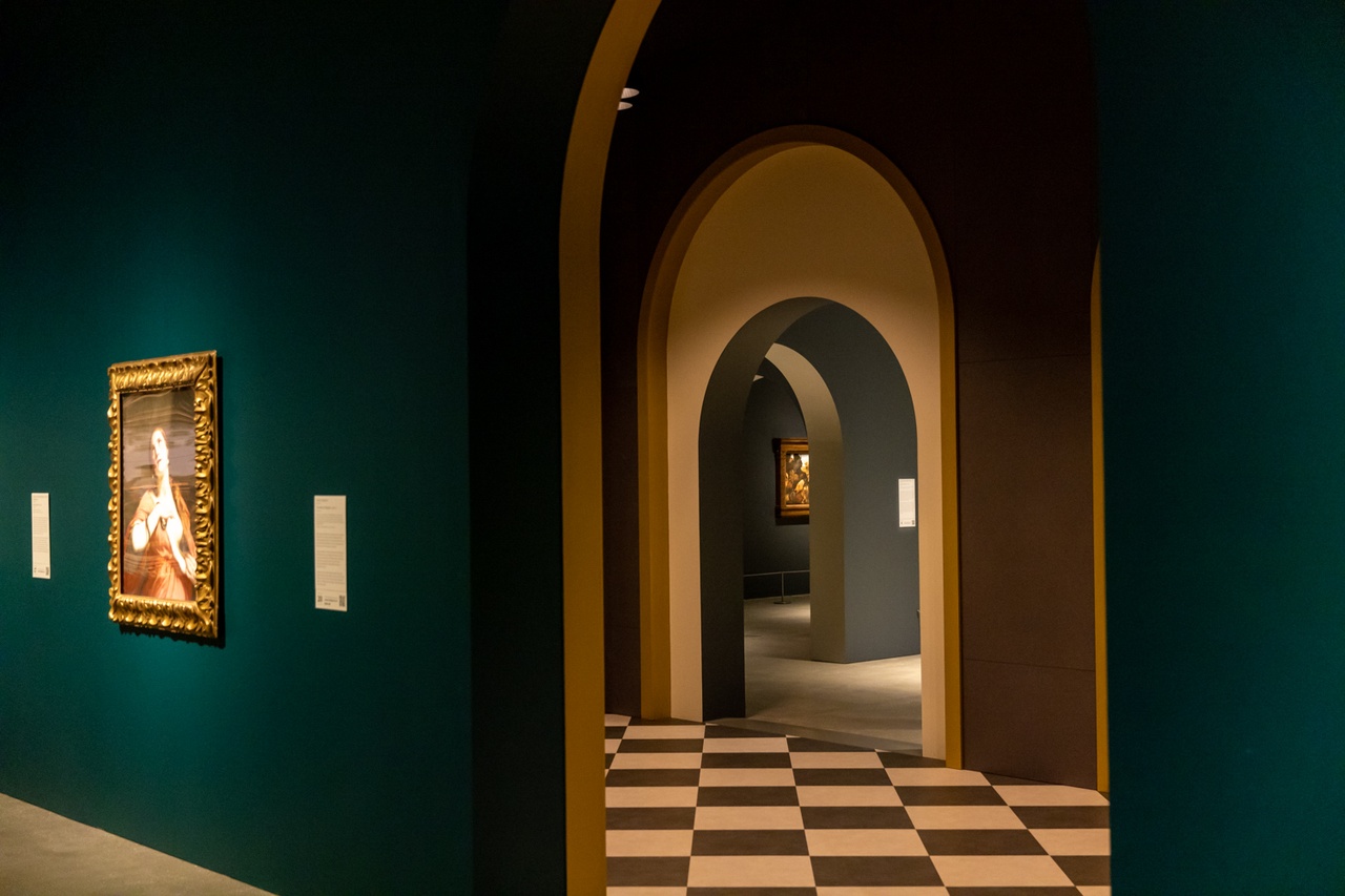 A hallway with green walls, archways and paintings hanging, with a check floor pattern.