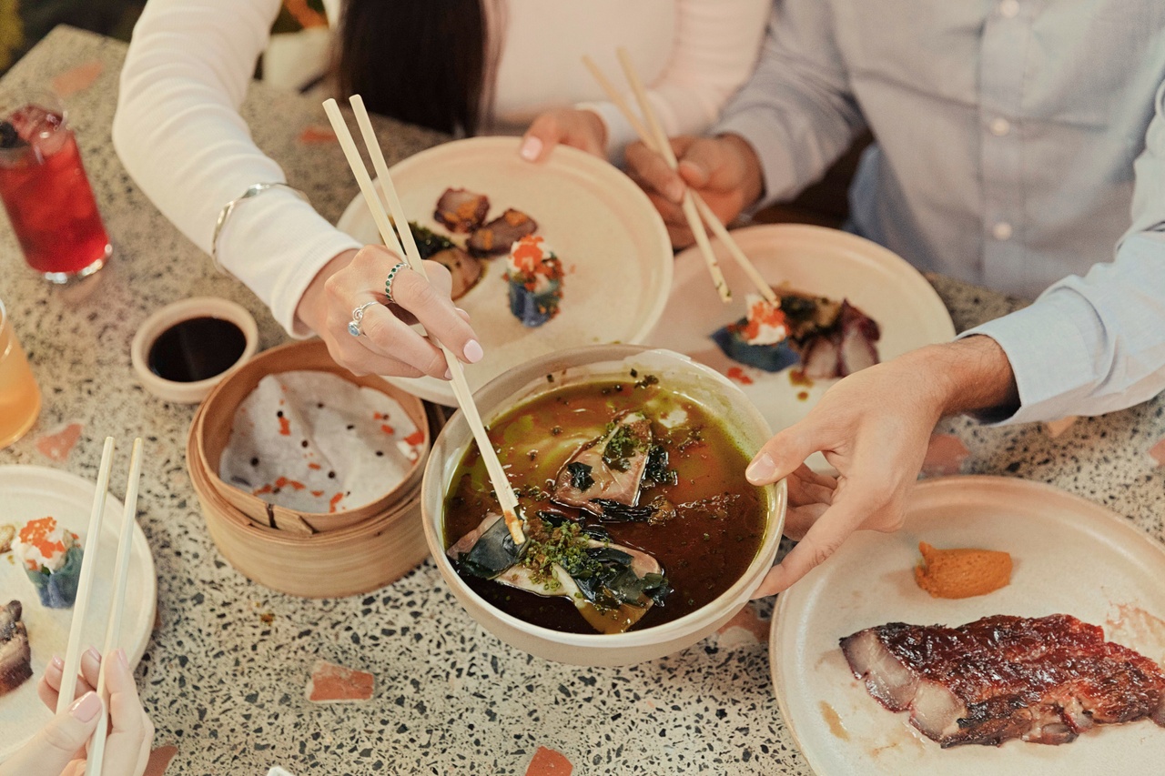 Hands reaching to food in bowls with chopsticks.