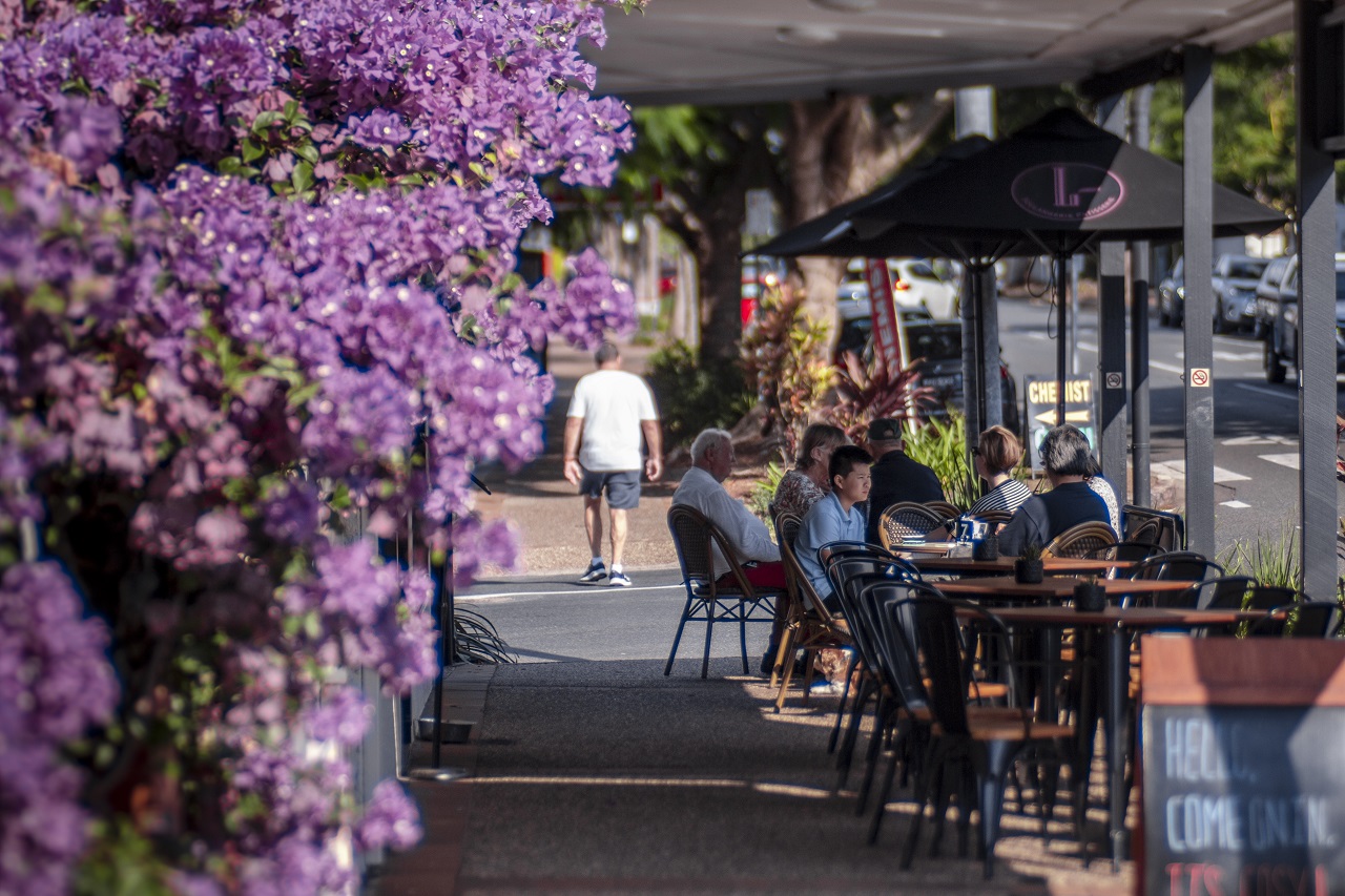 People dining at a sidewalk cafe in Ascot with a bougainvillea plant in the foreground.