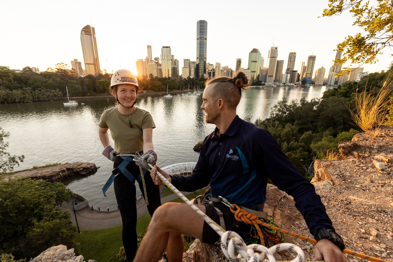 A woman starting to abseil down a cliff, being helped by an instructor, with Brisbane City in the background.