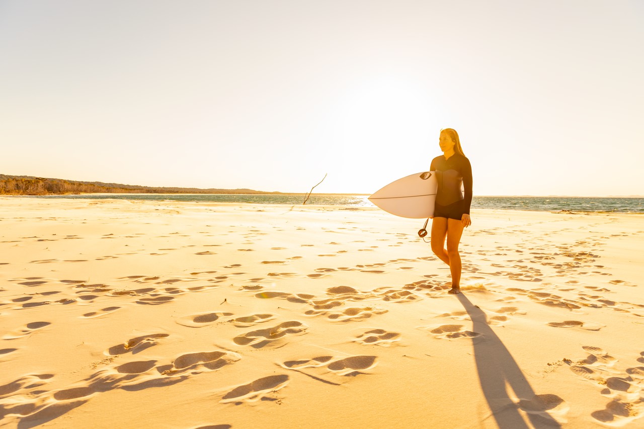 A woman walking on the beach with surfing board at North Stradbroke Island/Minjerribah